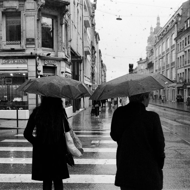 Black and white photo of people with umbrellas crossing a wet city street.