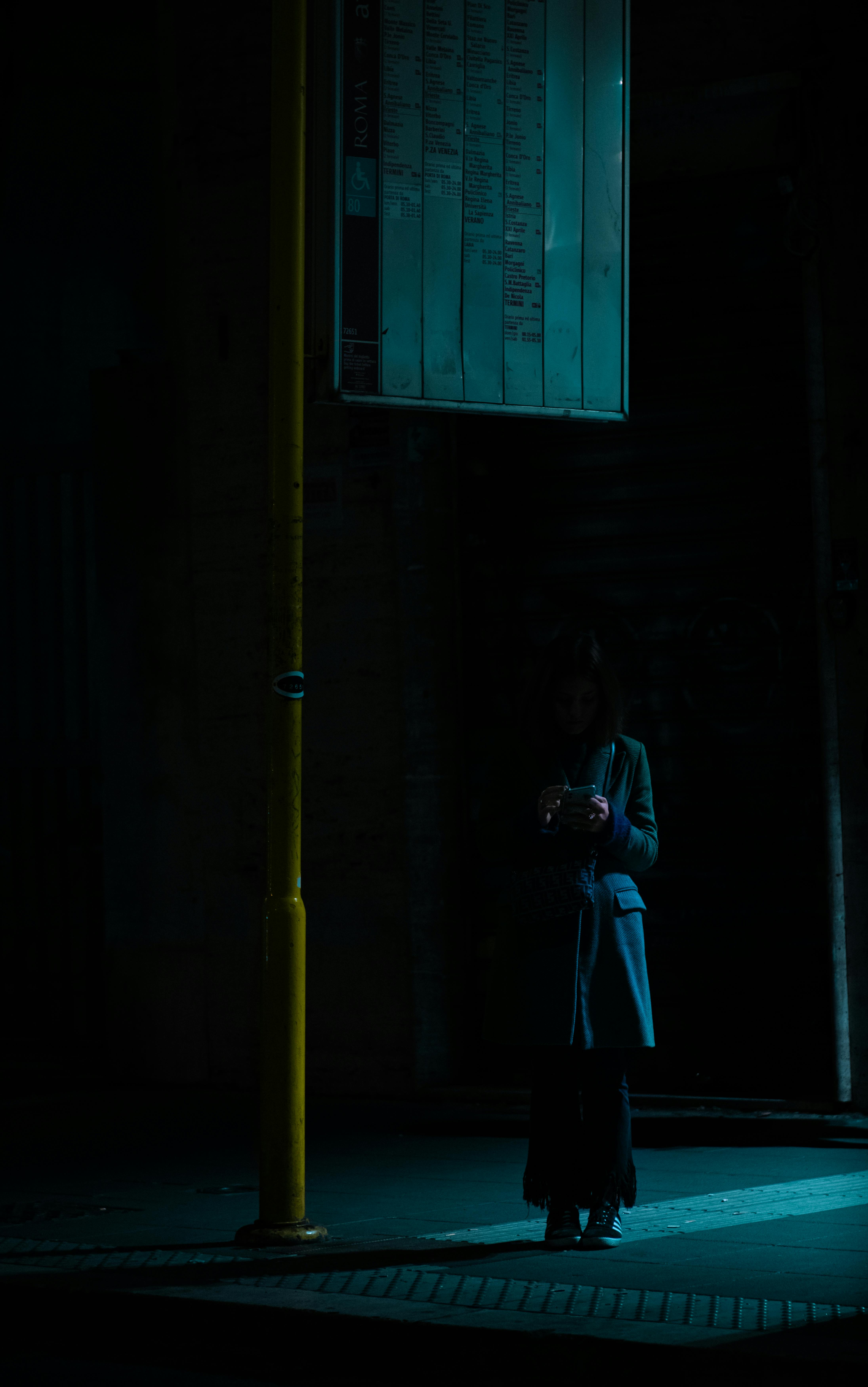 A solitary woman stands under a streetlight using her smartphone, creating a moody scene on a dark urban street.