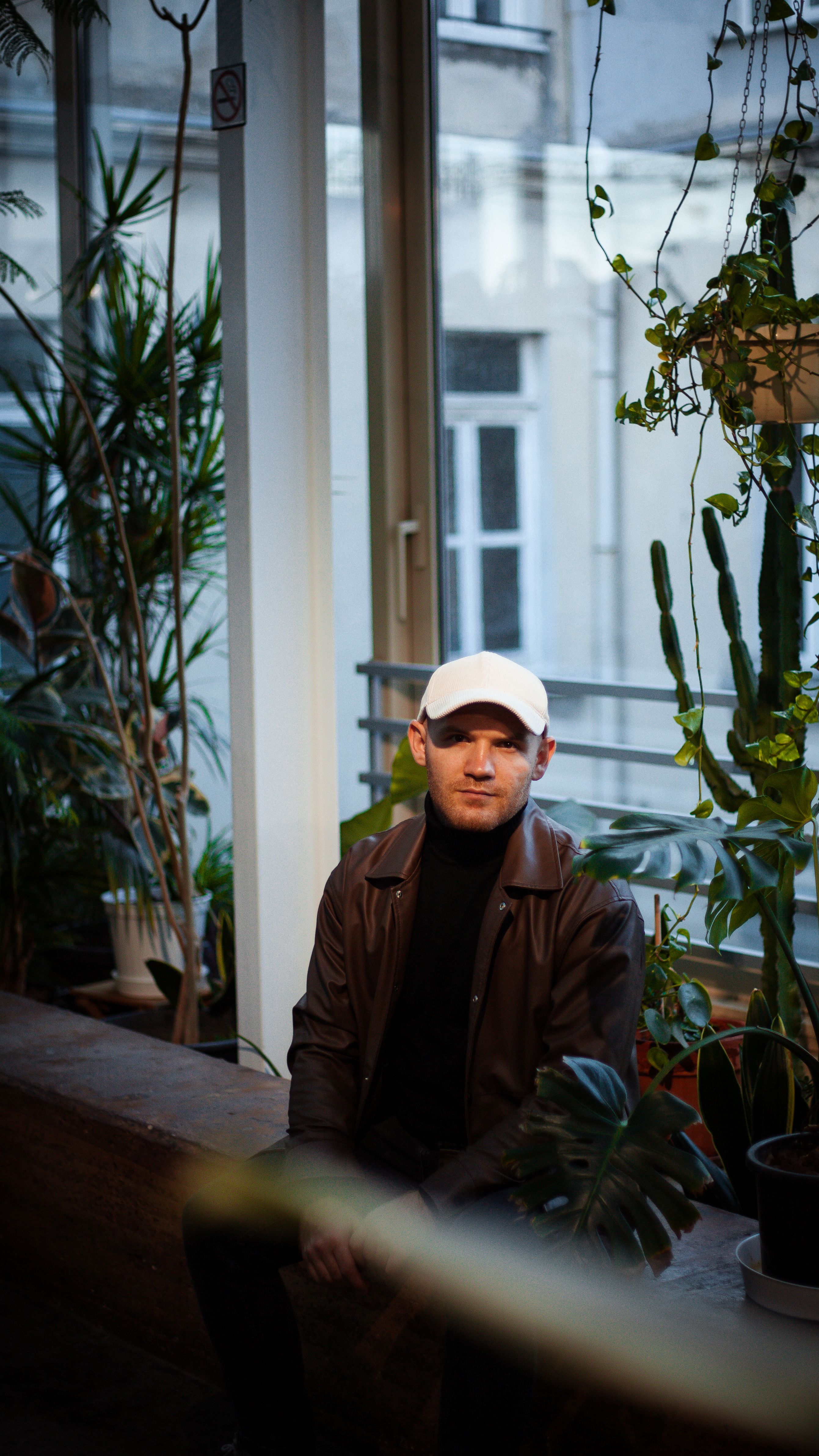 A man wearing a leather jacket and white cap sits in a stylish indoor garden setting.