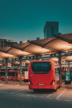 A vibrant red bus parked at a city bus station during evening hours, under illuminated canopies.