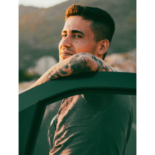 Free Side view of a tattooed man with an eyebrow piercing leaning on a car outdoors. Stock Photo