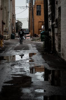 A dark, moody alleyway after rain featuring a solitary figure and puddle reflections.