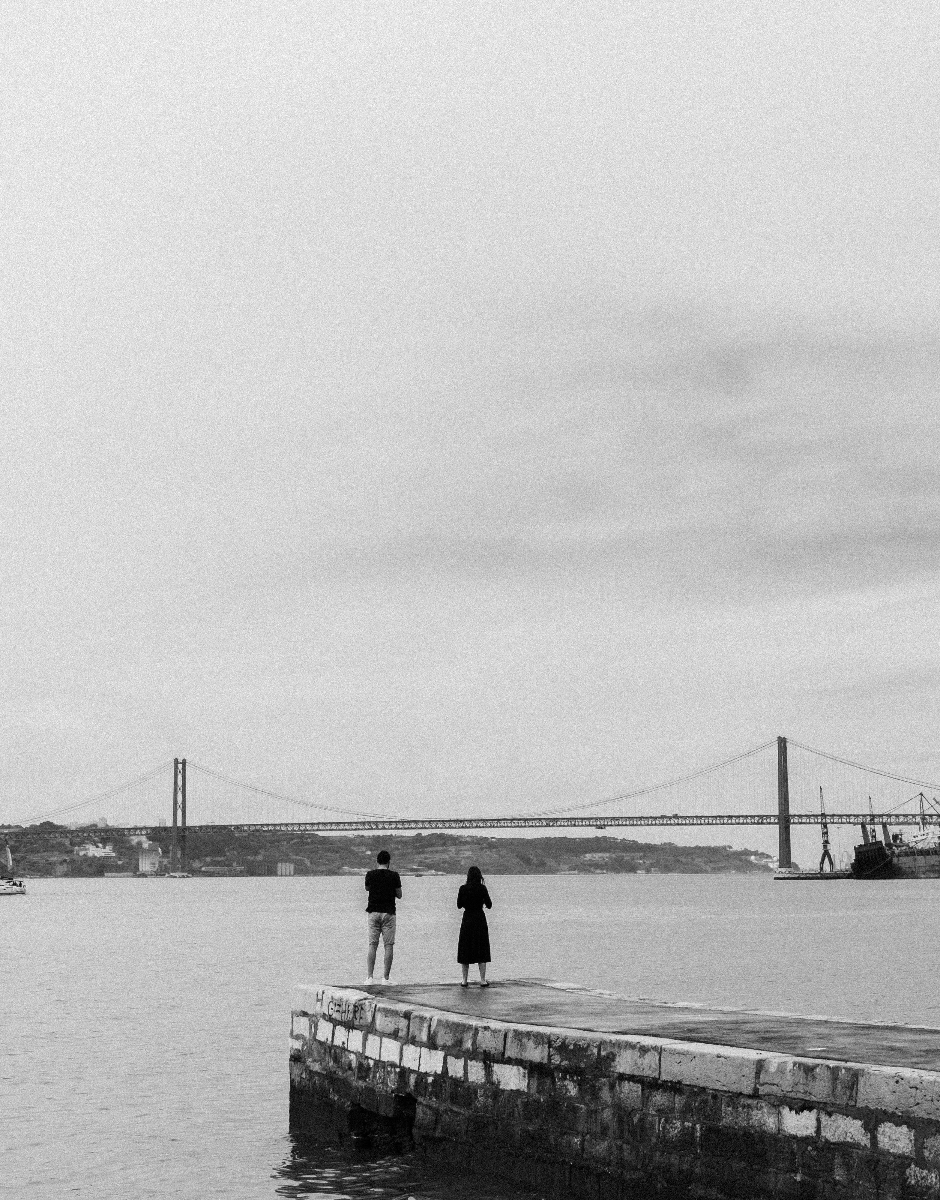 People standing on a dock in Porto with a suspension bridge in the background, captured in black and white.