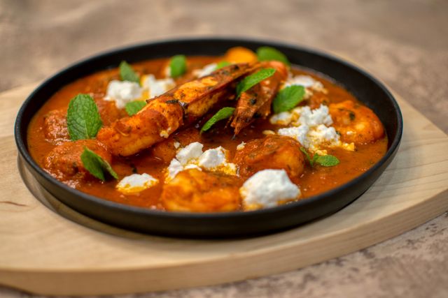 Close-up of a prawn curry garnished with cheese and fresh mint leaves, served in a black bowl.