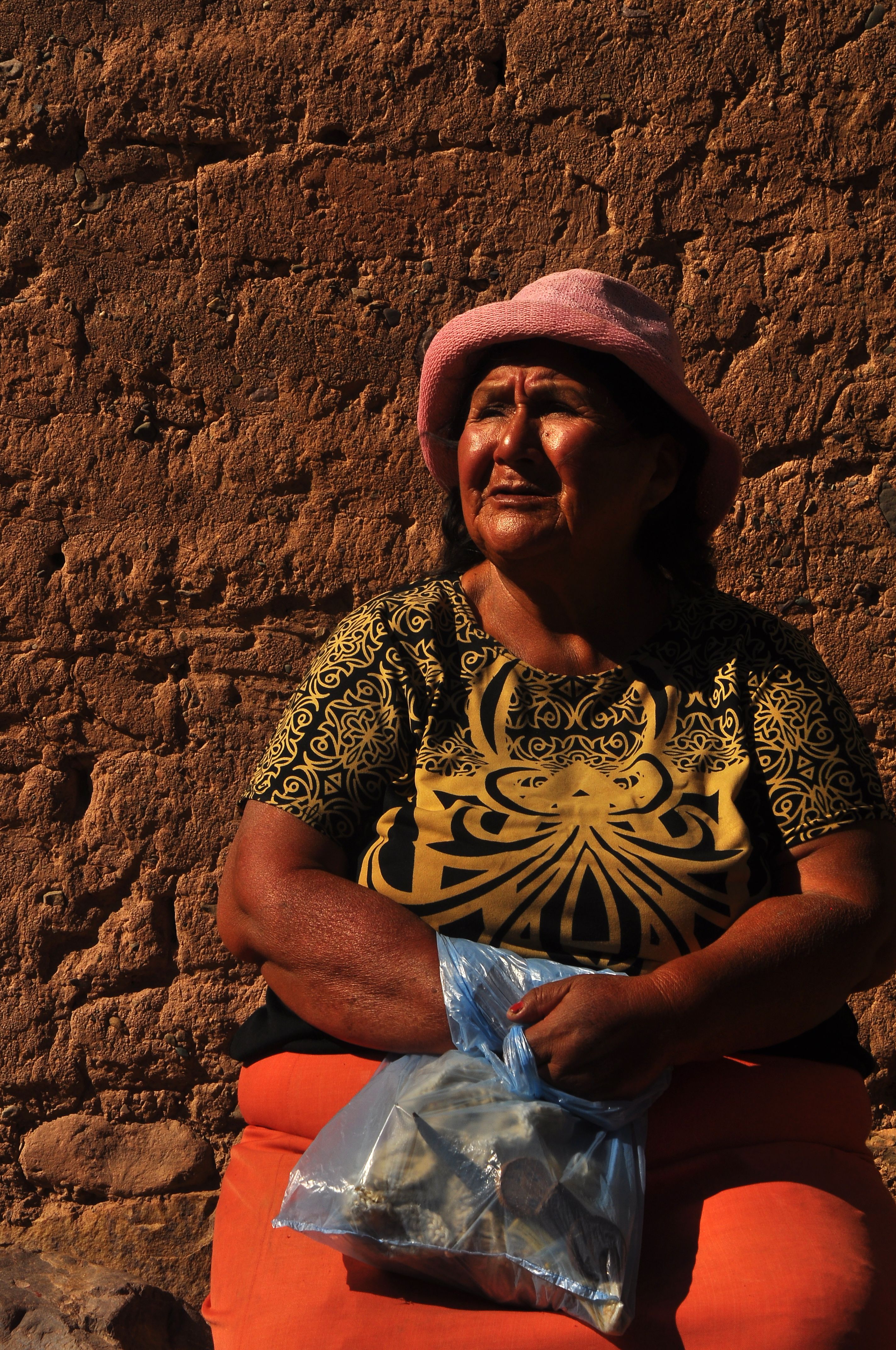 Elderly woman in patterned shirt and pink bucket hat sits holding a plastic bag against a textured wall.