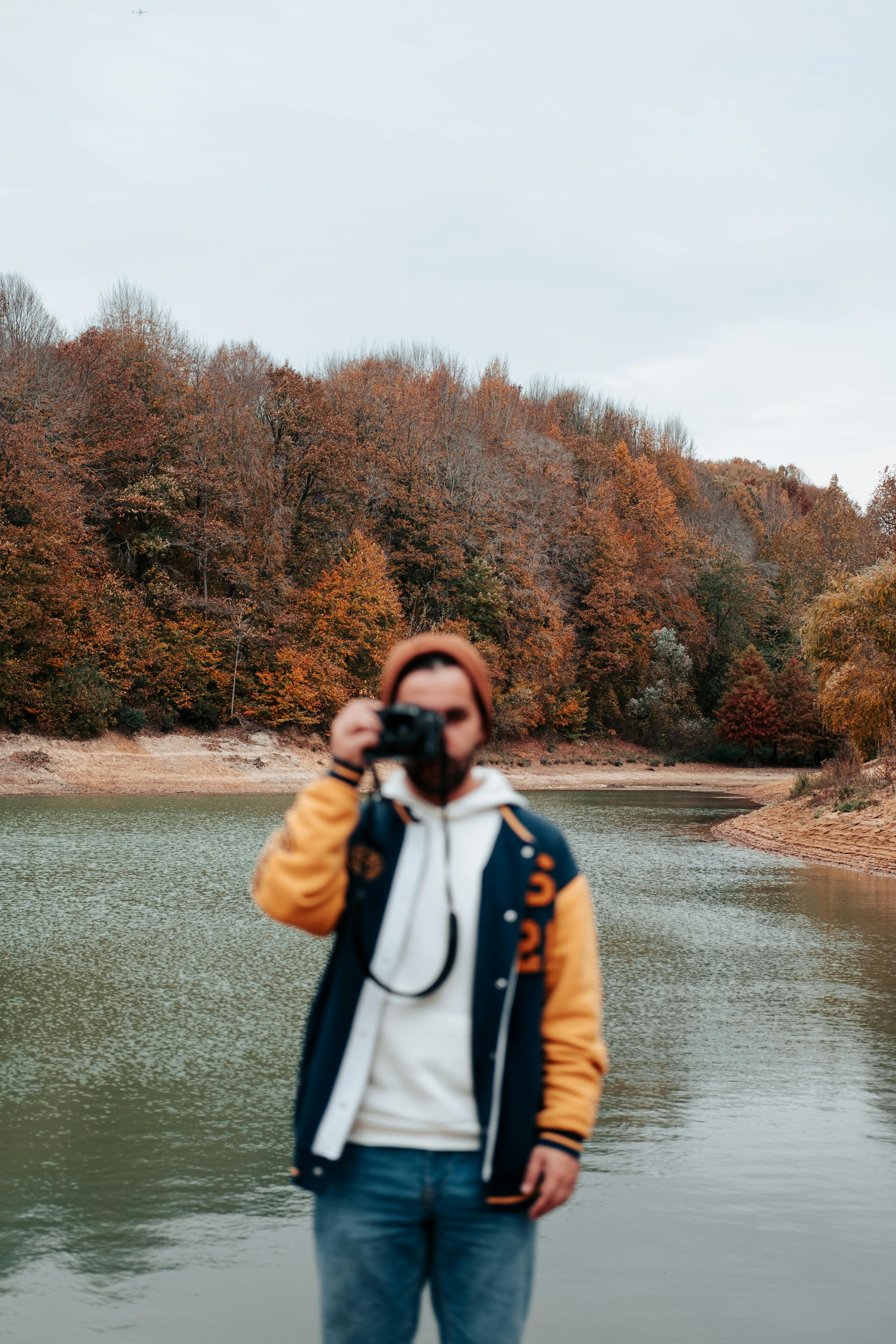 A man taking photos by a river with colorful autumn foliage in the background.