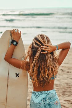 Blonde woman with surfboard standing on sunny beach, enjoying summer vibe.