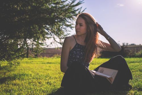 Free A woman sits in a grassy field reading a book under the sun, radiating calm and leisure. Stock Photo