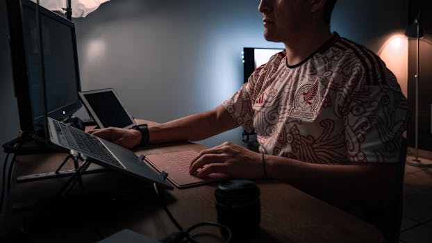 Man working on desktop computer and laptop in a dimly lit office setting.