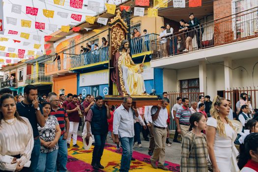 A vibrant street festival procession with a religious figure in Alto Lucero, Veracruz, Mexico, featuring colorful decorations and a large crowd.