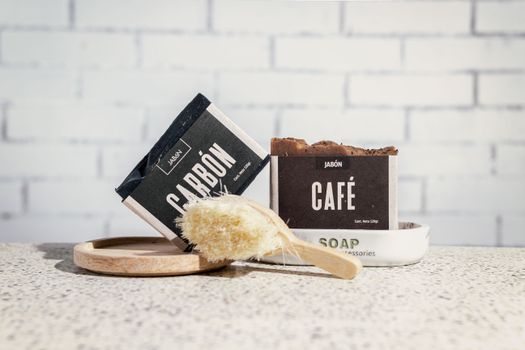 A close-up of artisanal coffee and charcoal soaps with a bath brush against a white brick wall. - Fotografer Sidoarjo