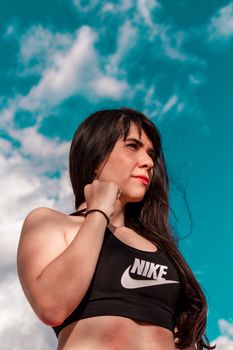 A woman in a black sports bra stands confidently against a bright blue sky with clouds.