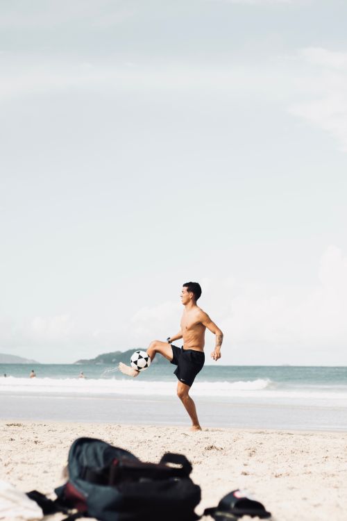 Free A man in swim trunks playing football on a sandy beach under a clear sky. Stock Photo