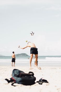 A group of shirtless men playing soccer on a sunny beach with ocean waves in the background.