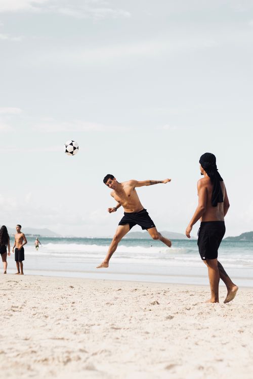 Free Shirtless men playing soccer on a sandy beach, enjoying a sunny day by the ocean. Stock Photo