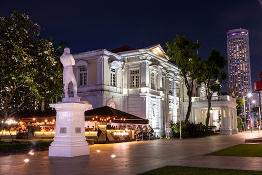 An illuminated historic building with a statue and city lights at night in Singapore, capturing urban charm.