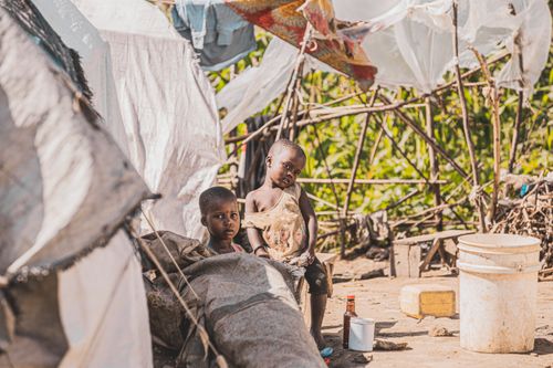Free Two children sitting by makeshift shelters in a rural village, surrounded by nature. Stock Photo