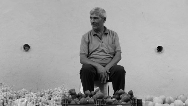 A monochrome portrait of an elderly man sitting by his fruit stall in a market.