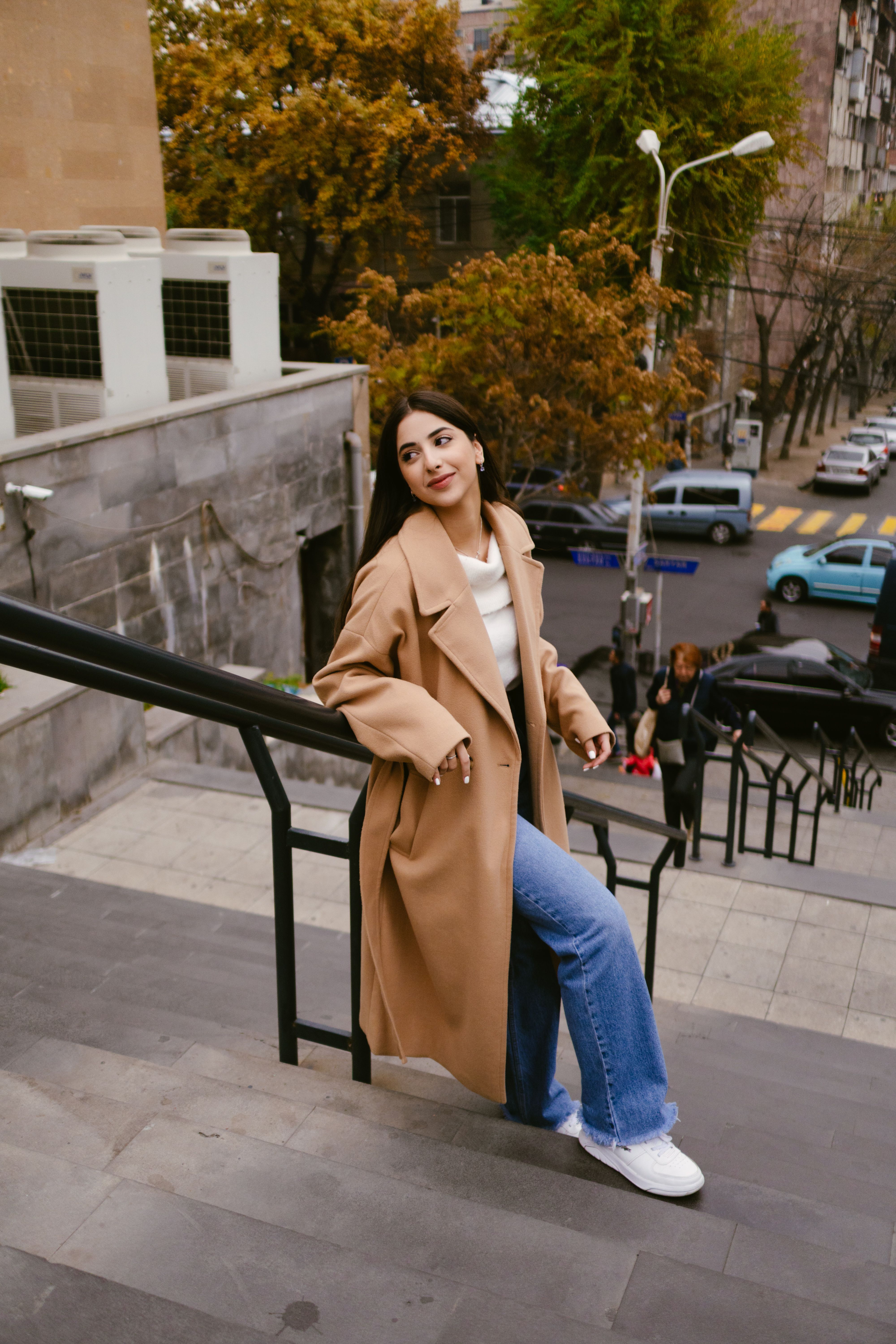 Fashion-forward woman in a beige coat posing on city stairs in Yerevan, Armenia.