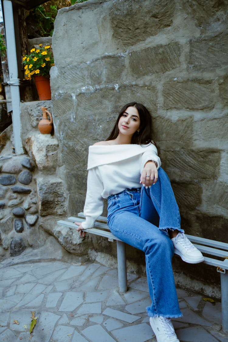 Woman In White Off Shoulder Sweater And Denim Pants Sitting On A Metal Bench