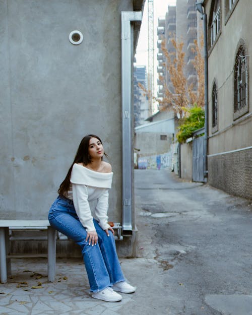 Woman in White Long Sleeve Shirt and Blue Denim Jeans Sitting on Concrete Bench