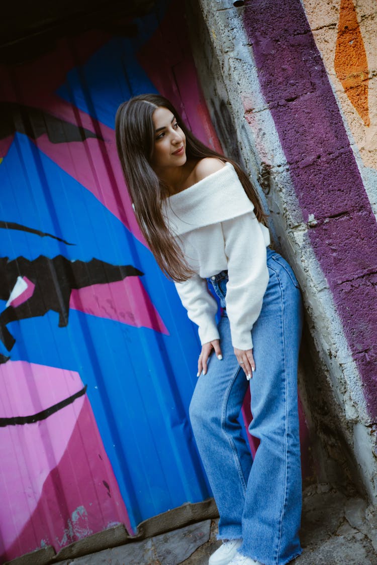 Young Woman In White Off Shoulder Sweater And Denim Jeans Leaning Against A Graffiti Wall