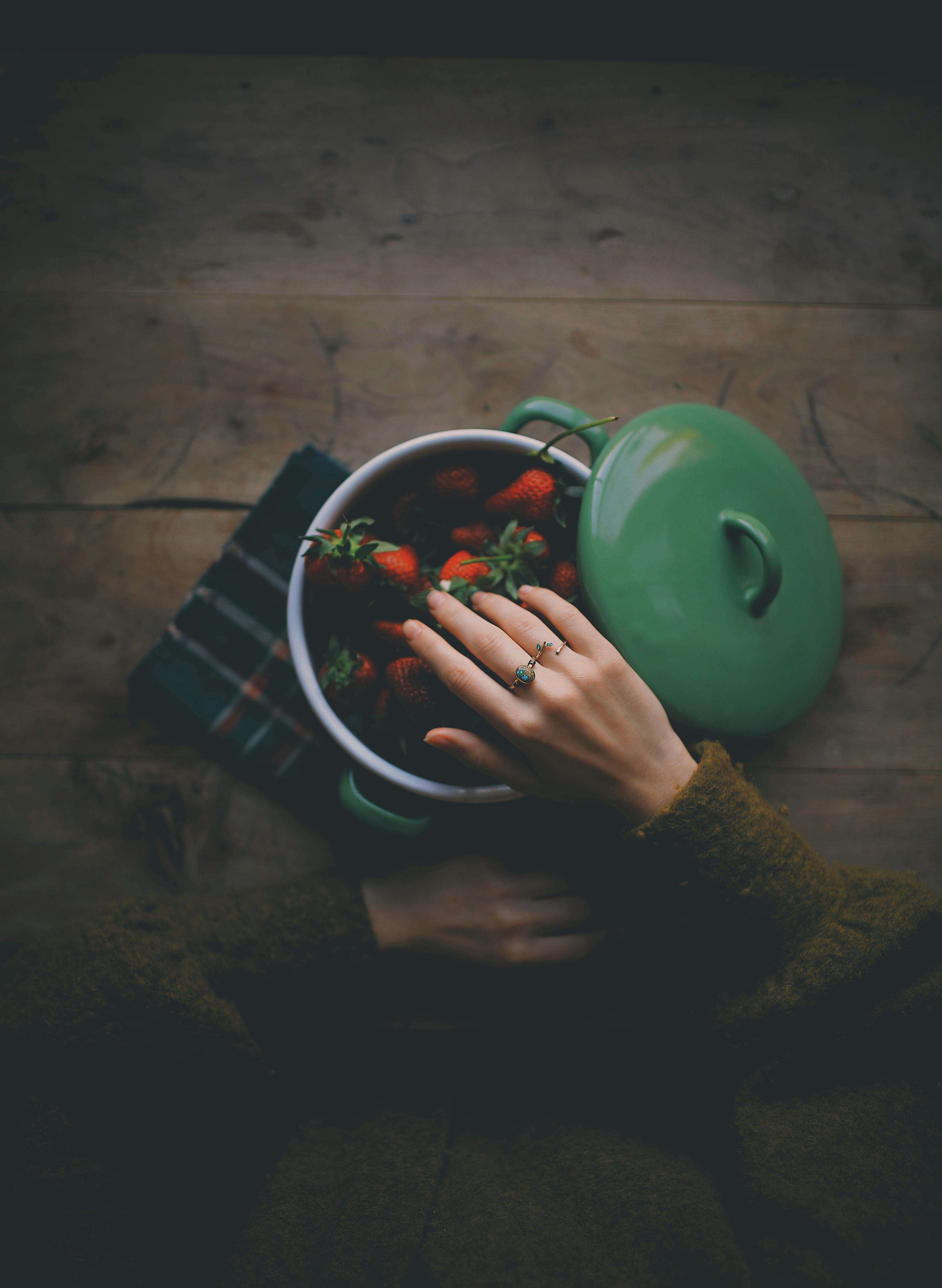 A hand reaching for strawberries in a green pot on a wooden table.