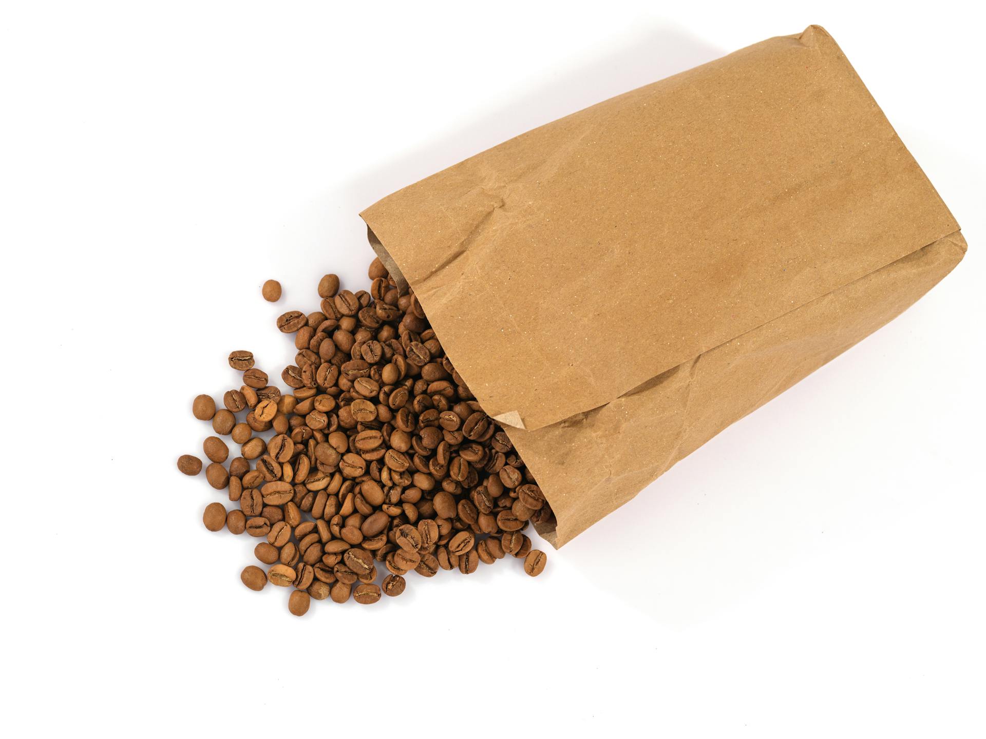 Brown paper bag with aromatic roasted coffee beans spilled on white background.