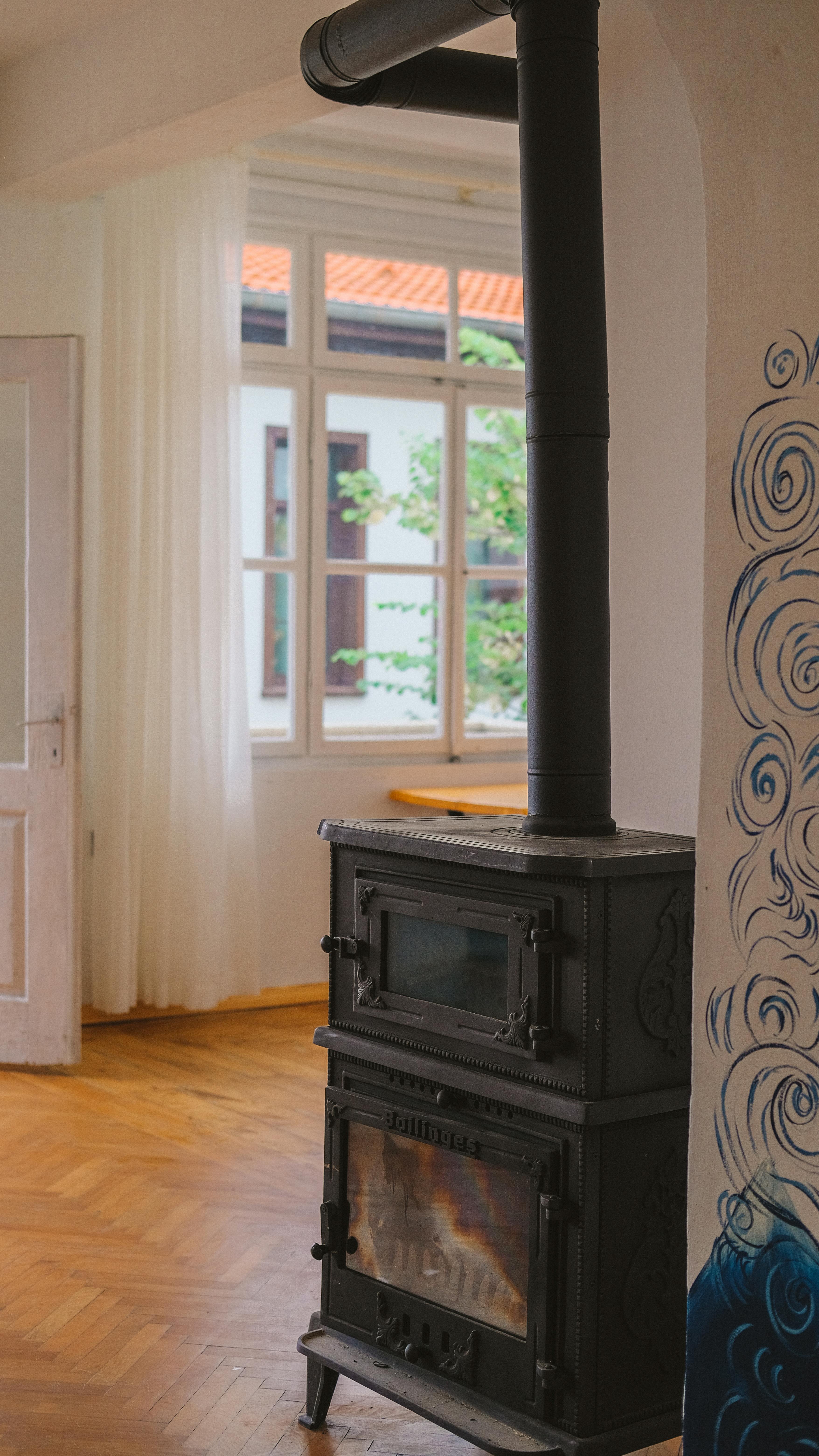 A black cast iron stove with a chimney in a room with a window and hardwood floors.