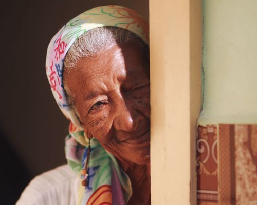 Free A serene elderly woman with gray hair smiles warmly, wearing a colorful headscarf, enjoying the sunlight indoors. Stock Photo