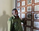 An older military veteran in uniform, stands by a wall of historical black and white portraits indoors.