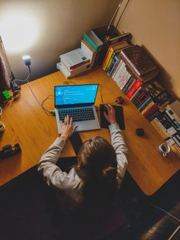 Teenage girl using a laptop on a cluttered desk with books, studying in a cozy room.