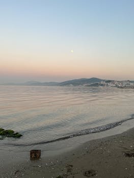 Peaceful sunrise at the beach with calm waters and distant mountains under a pastel sky.