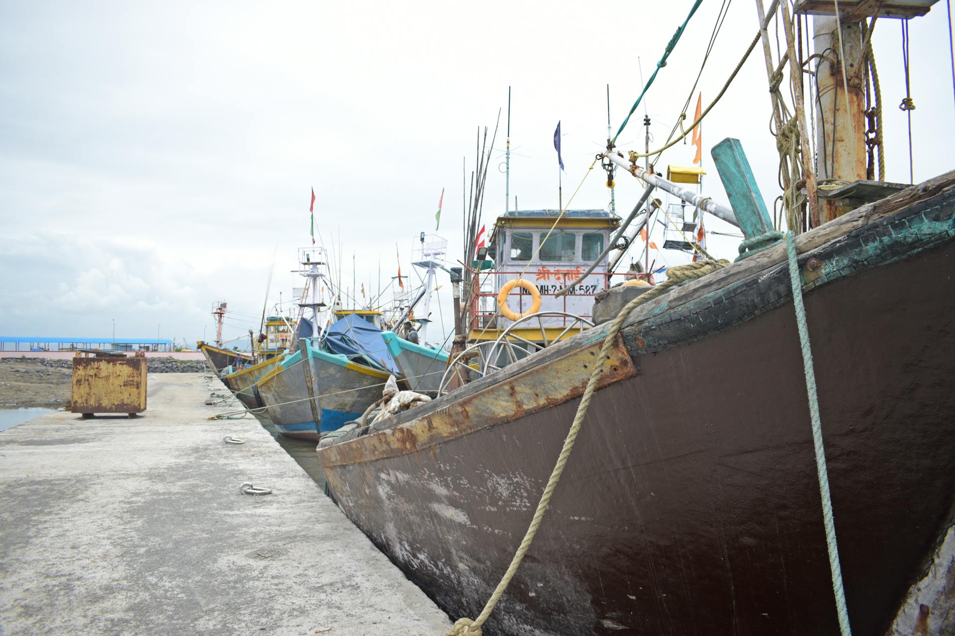 A row of weathered fishing boats docked at a harbor's concrete pier under an overcast sky.