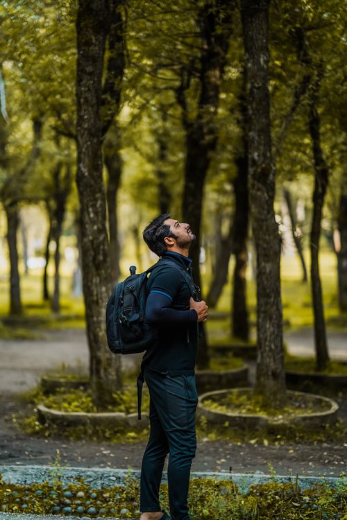 Free Man with backpack exploring a lush green forest, looking upward thoughtfully. Stock Photo
