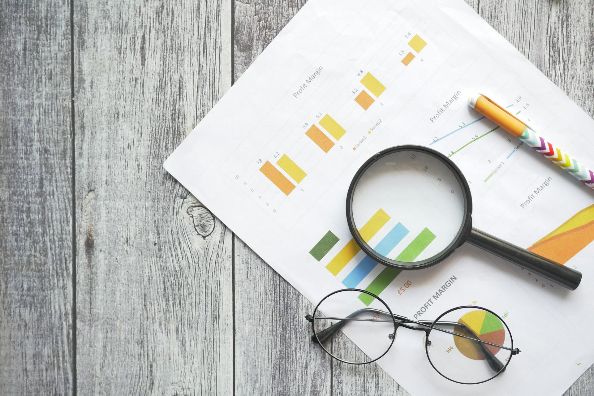 Overhead view of financial charts, a magnifying glass, ballpen, and eyeglasses on a wooden surface.