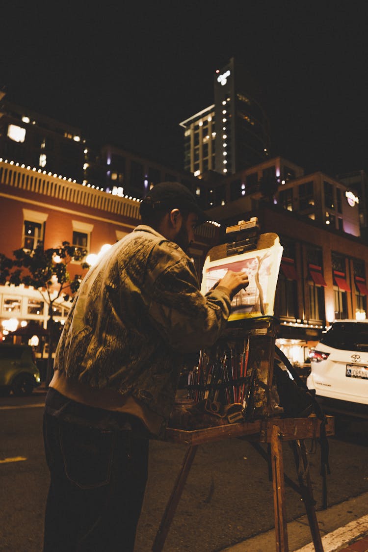 Man Standing On The Street And Painting A Picture At Night 