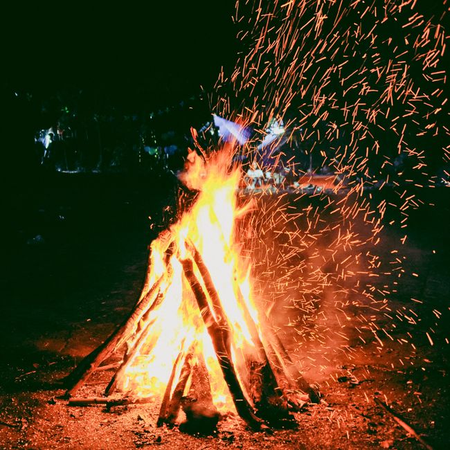 High flames and sparks fly from a vibrant outdoor campfire at night, creating a warm and lively scene.