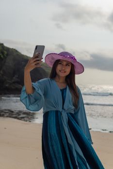A woman in a flowy blue dress and purple hat takes a selfie on a scenic beach in Pacitan.