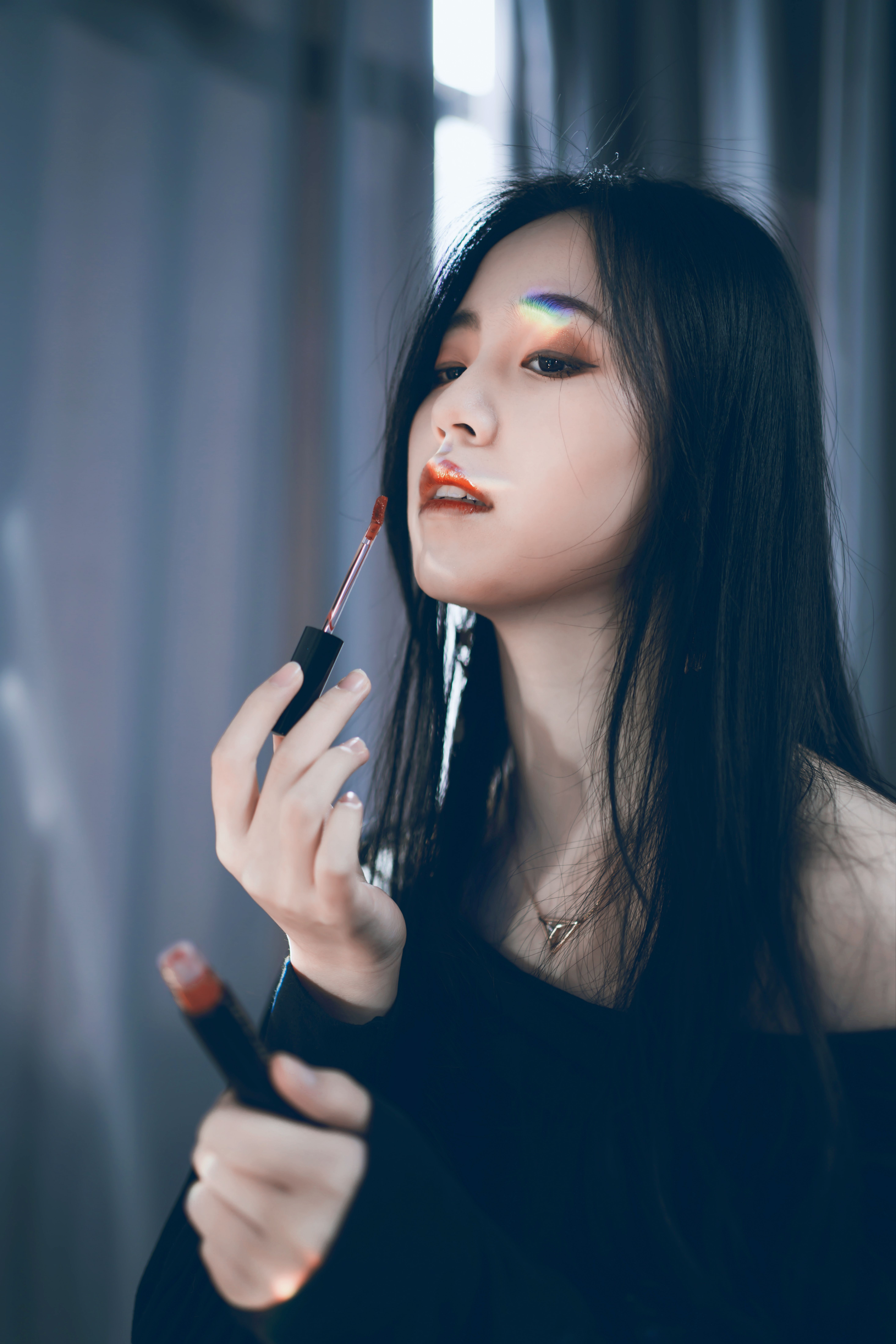 Stylish Asian woman in black applies lipstick indoors with soft lighting and a touch of rainbow.
