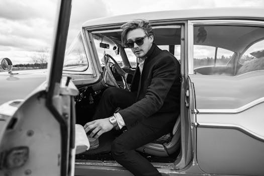 A man in sunglasses sits in a vintage car, captured in classic black and white style.