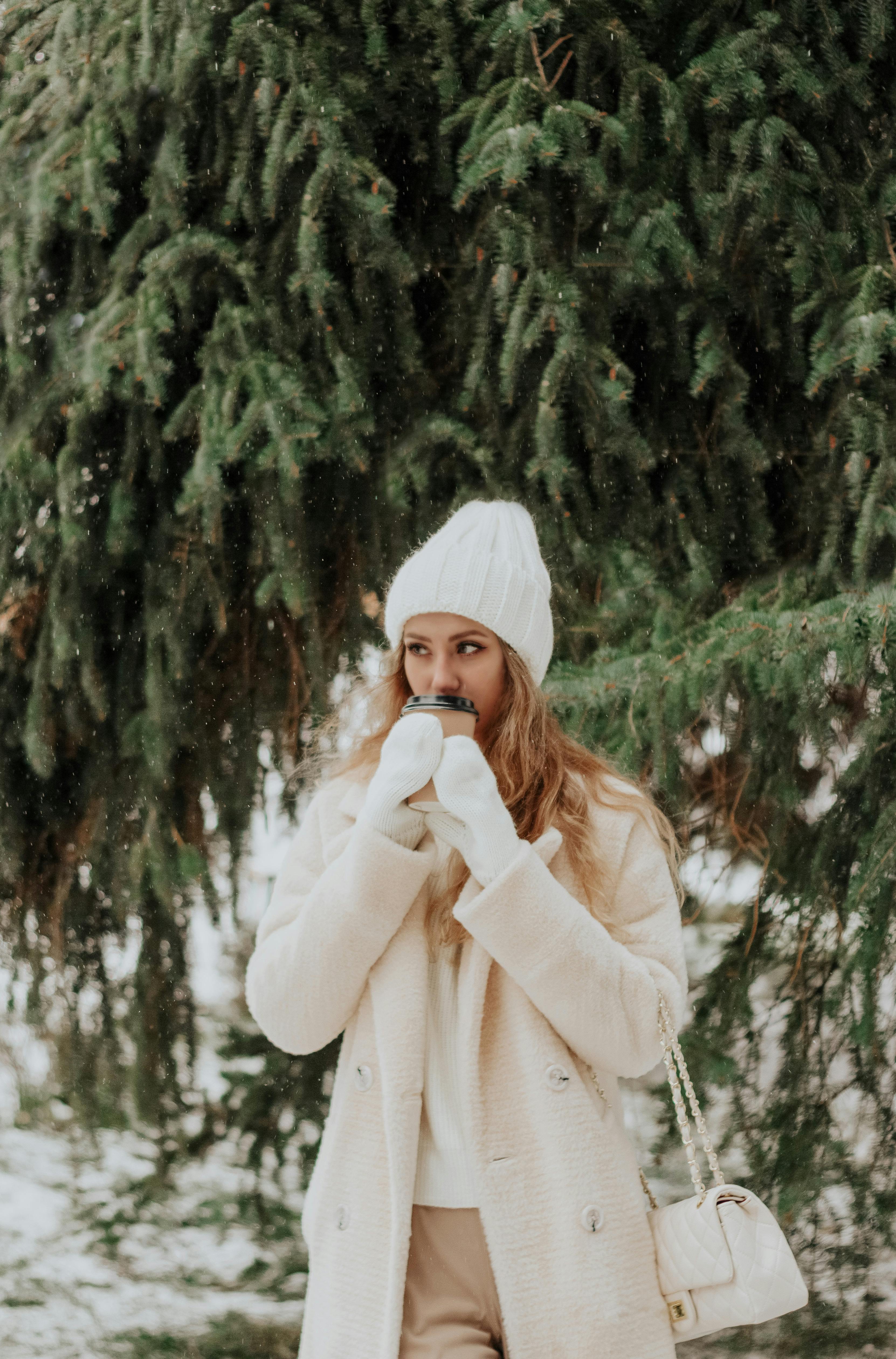 Stylish woman in winter coat and hat sipping hot drink outdoors with evergreen trees.