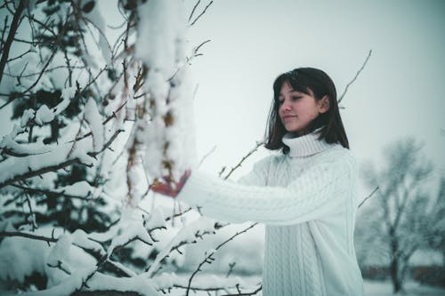 A Woman in White Knitted Sweater Standing Near the Snow Covered Tree