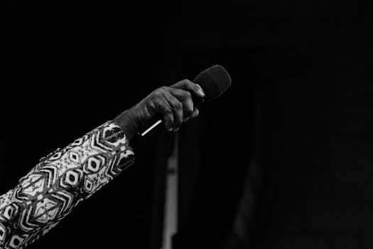 Artistic monochrome image depicting a hand in patterned clothing holding a microphone against a dark background.