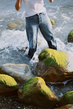 A person in jeans stands in ocean waves with moss-covered rocks around them.
