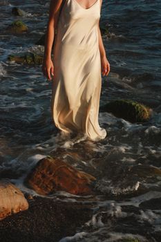 A woman in a flowing dress strolls along a rocky coast with gentle waves.