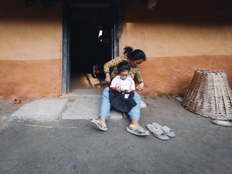 A mother assists her child with school uniform, sitting outside a rustic house.