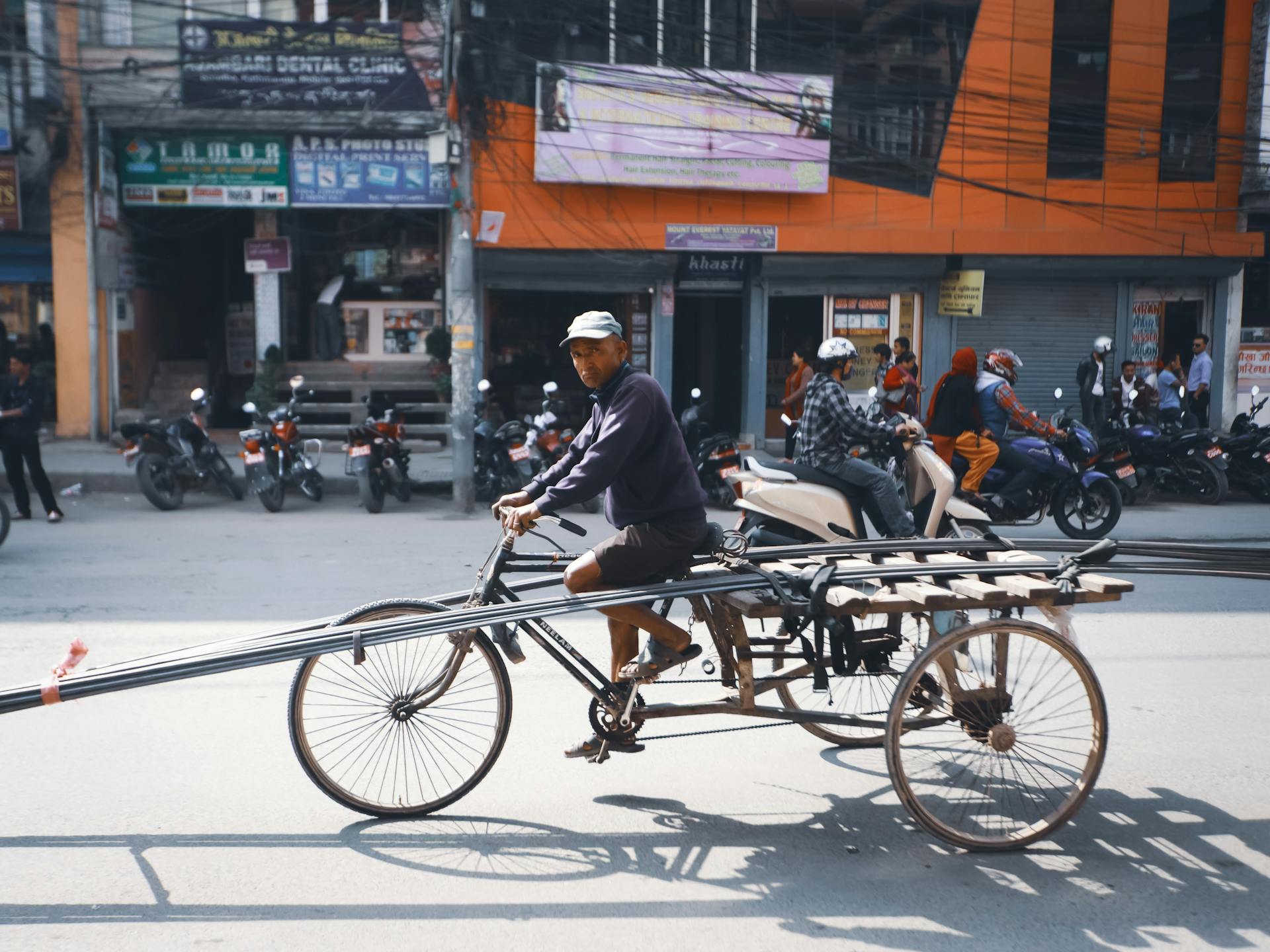 Man on a tricycle carrying long beams through a lively city street with motor scooters passing by.