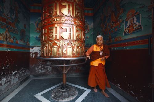 Free A senior monk stands by a large prayer wheel in an ornate temple, showcasing traditional Buddhism culture. Stock Photo