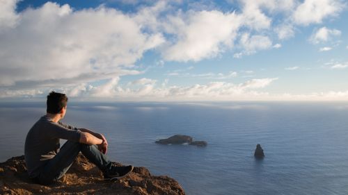 Free A man sits on a cliff in Hanga Roa, Chile, overlooking the ocean under a blue sky with white clouds. Stock Photo